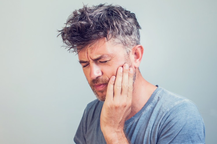 Middle-aged man wearing a gray shirt cringes in pain due to a toothache dental emergency