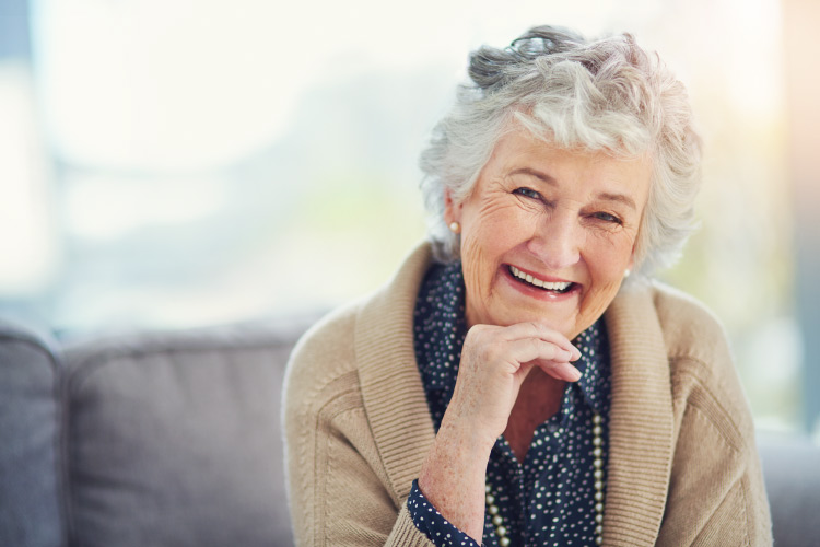 Cute older gray haired lady wearing a tan cardigan and smiling with her hand under her chin