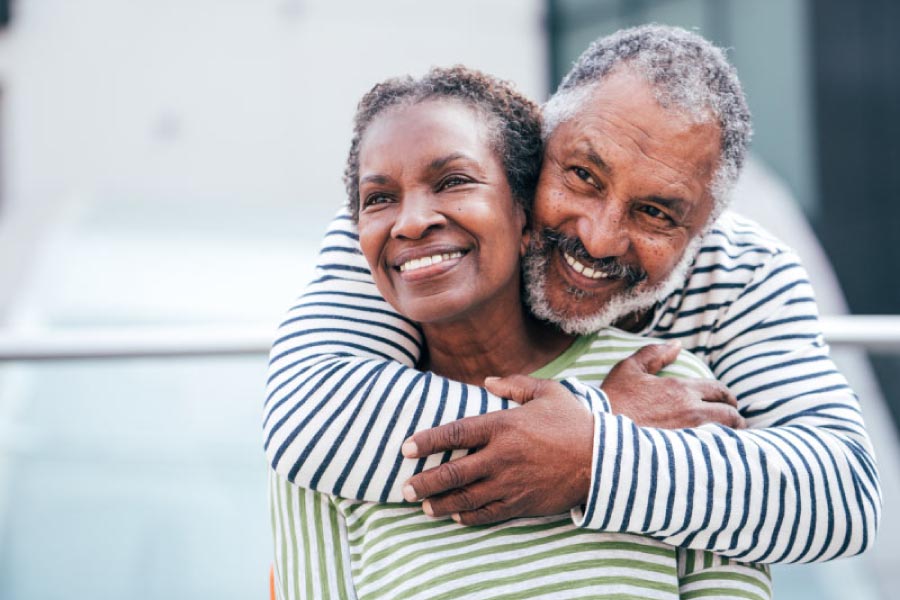 Smiling older black couple with the man standing behind the woman and giving her a hug